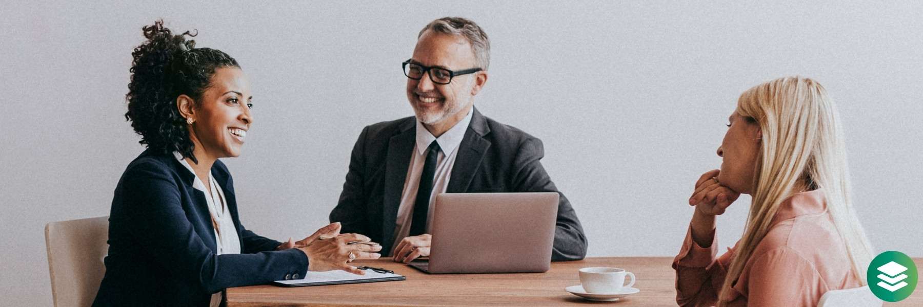 Three business professionals sitting at a table having a conversation while one takes notes and one has their laptop open