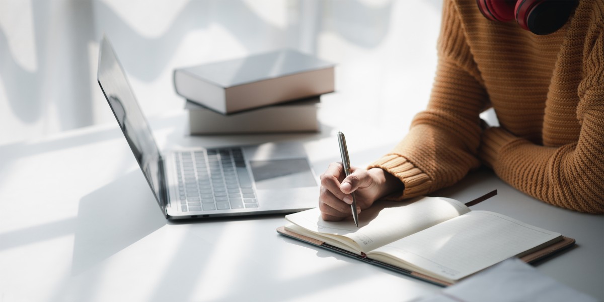 A person at a table taking notes in a notebook next to their open laptop and stacked books