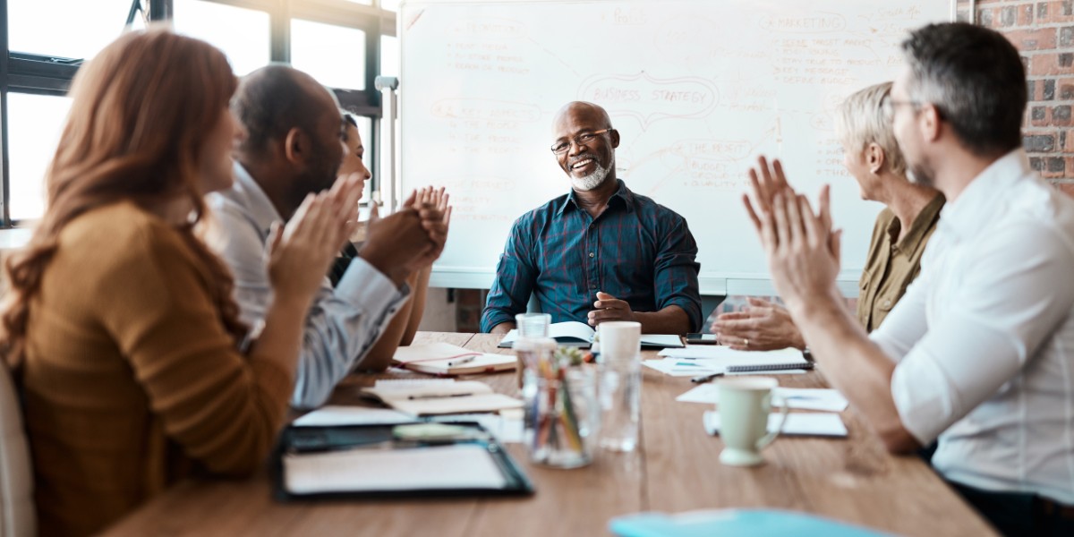 A group of business professionals sitting around a meeting table clapping for the leader sitting at the head of the table