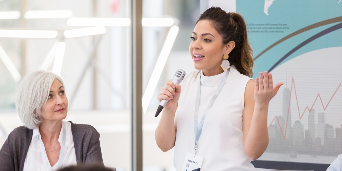 A speaker at an event holding a microphone and speaking with a big smile on their face
