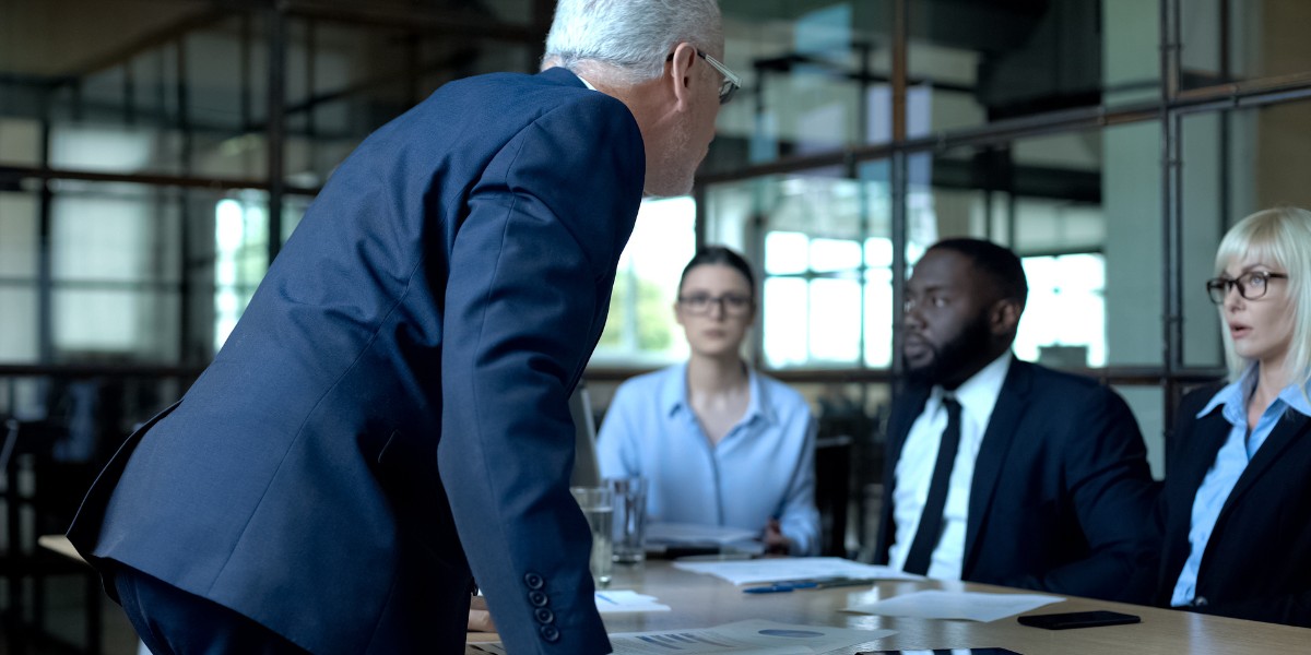 A person is leaning in at a conference table while three people on the other side of the table appear uncomfortable