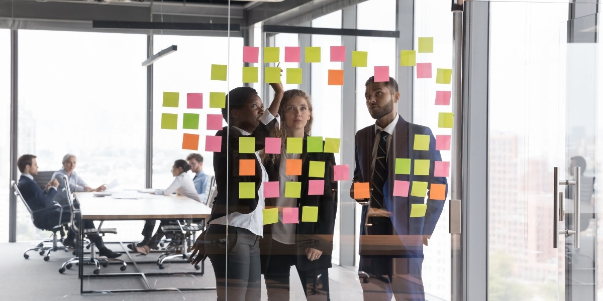 Three project team members are standing at a project Kanban board discussing items on the sticky notes while the rest of the project team chats at a table behind them