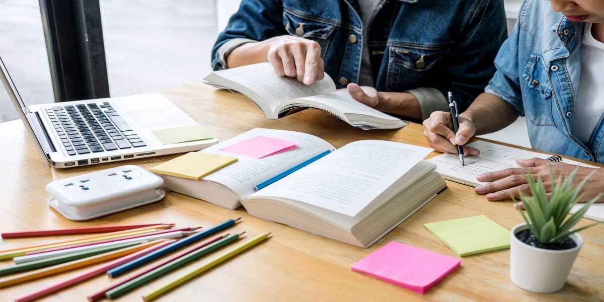 Two people sitting at a table studying and writing notes with open books and a laptop on the table